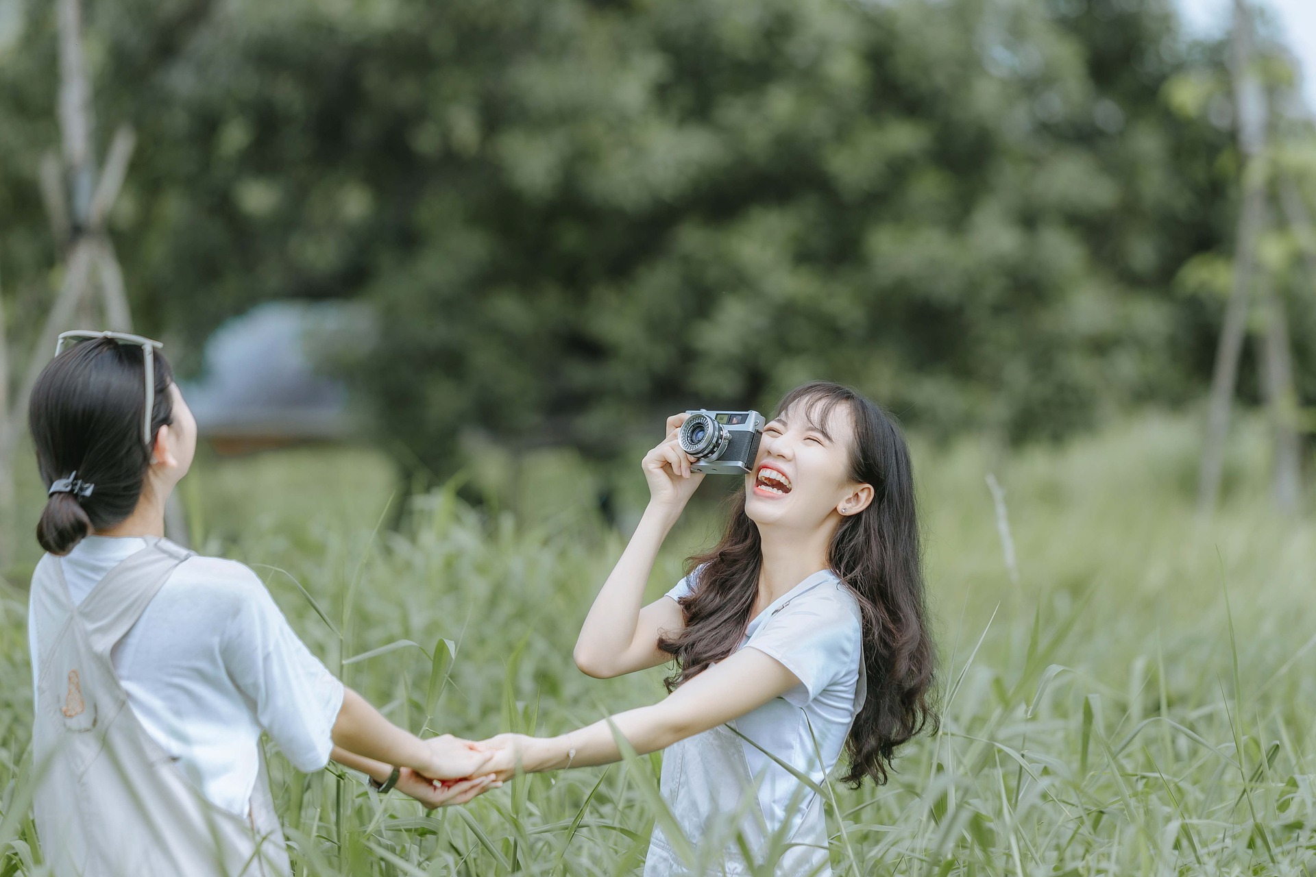 Two young women in a green field, laughing and holding hands, one has a vintage camera in her hand Two young women in a green field, laughing and holding hands, one has a vintage camera in her hand
